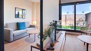A modern living room with a sofa, coffee table, and a dining area with chairs. There is a large window providing a view of the outdoors. -  Livensa Living Studios Madrid Alcobendas (Alcobendas)