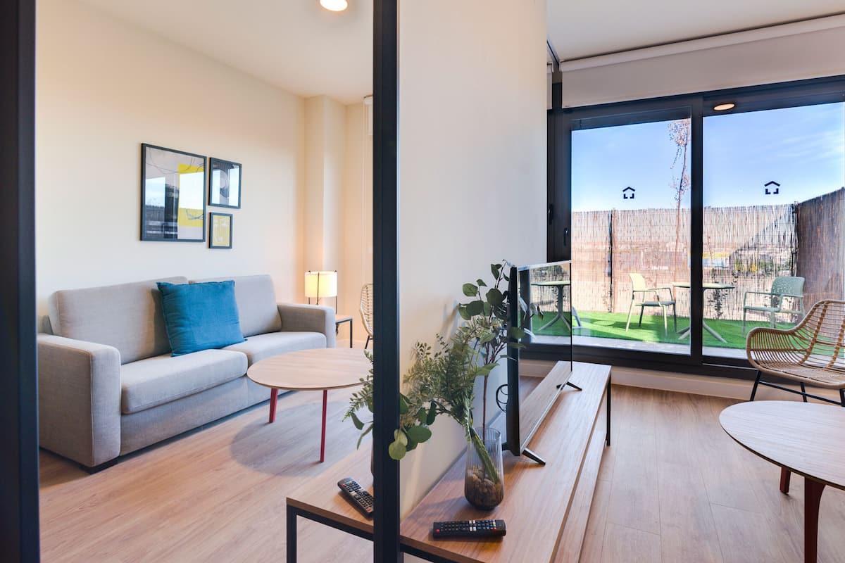 Un salon moderne avec un canapé, une table basse et un coin repas avec des chaises. Une grande fenêtre offre une vue sur l’extérieur.