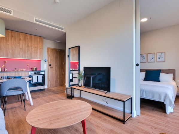Une chambre d’hôtel moderne avec un plancher en bois, un lit avec une literie blanche et turquoise, un téléviseur posé sur un meuble en bois, un bureau avec une chaise et, au fond, une petite cuisine.