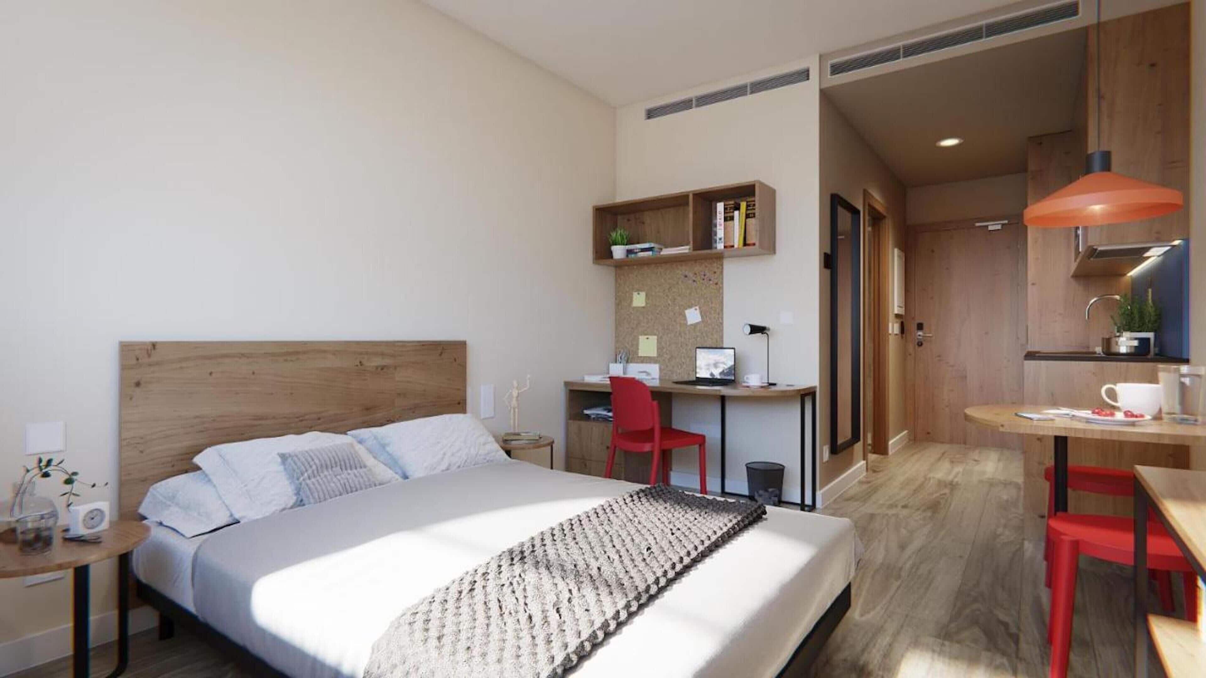 A modern hotel room with a wooden bed, a desk with a computer, a red chair, and a small kitchenette.