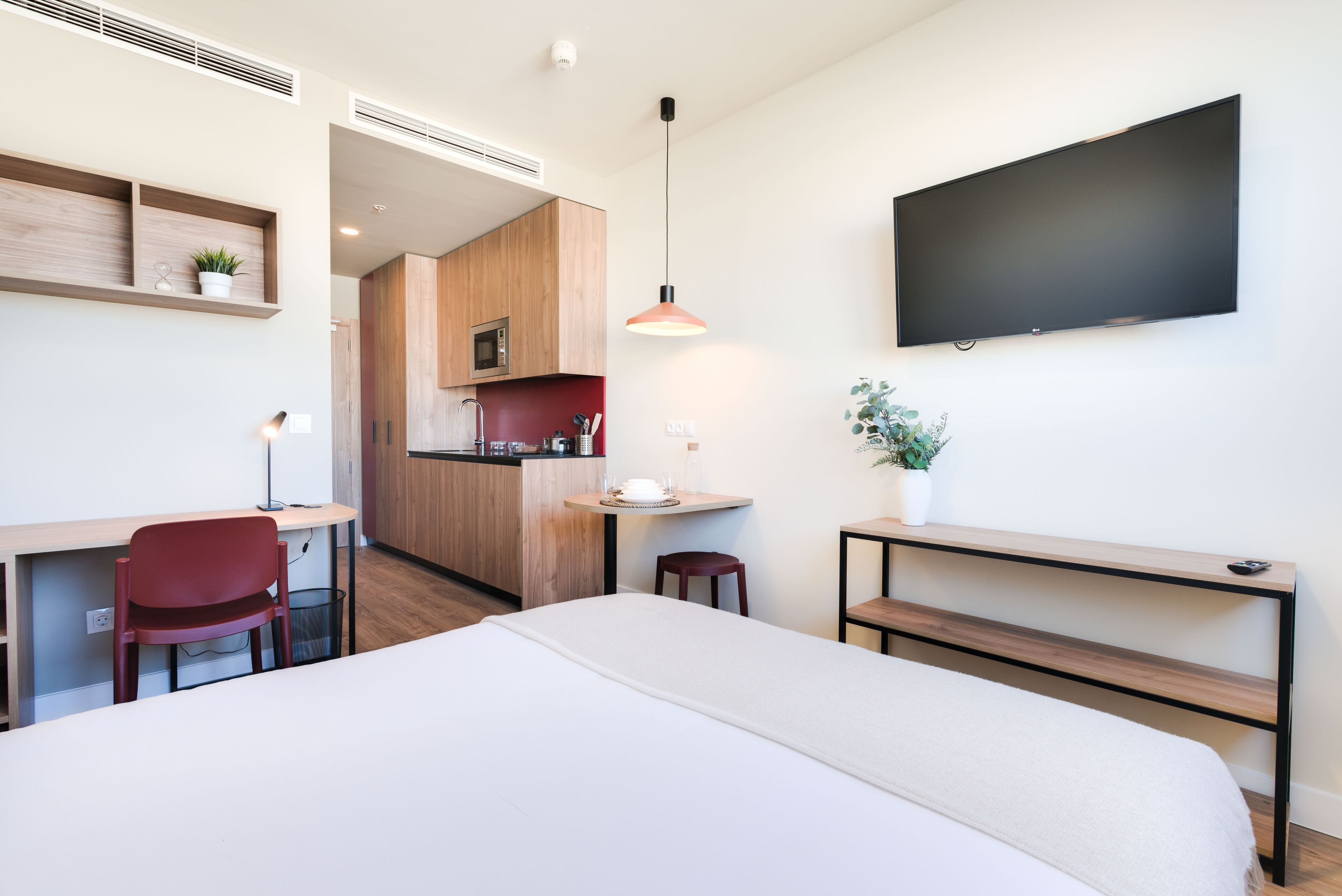 Una habitación de hotel moderna con una cama, un escritorio, una silla, un televisor montado en la pared y una pequeña cocina.