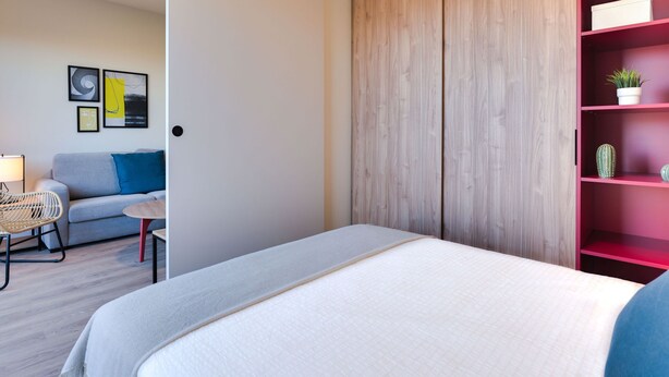 Une chambre à coucher moderne avec un lit, un canapé gris, un fauteuil en osier et une étagère rouge ornée de plantes.