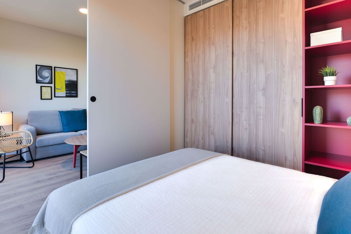 Une chambre à coucher moderne avec un lit, un canapé gris, un fauteuil en osier et une étagère rouge ornée de plantes.