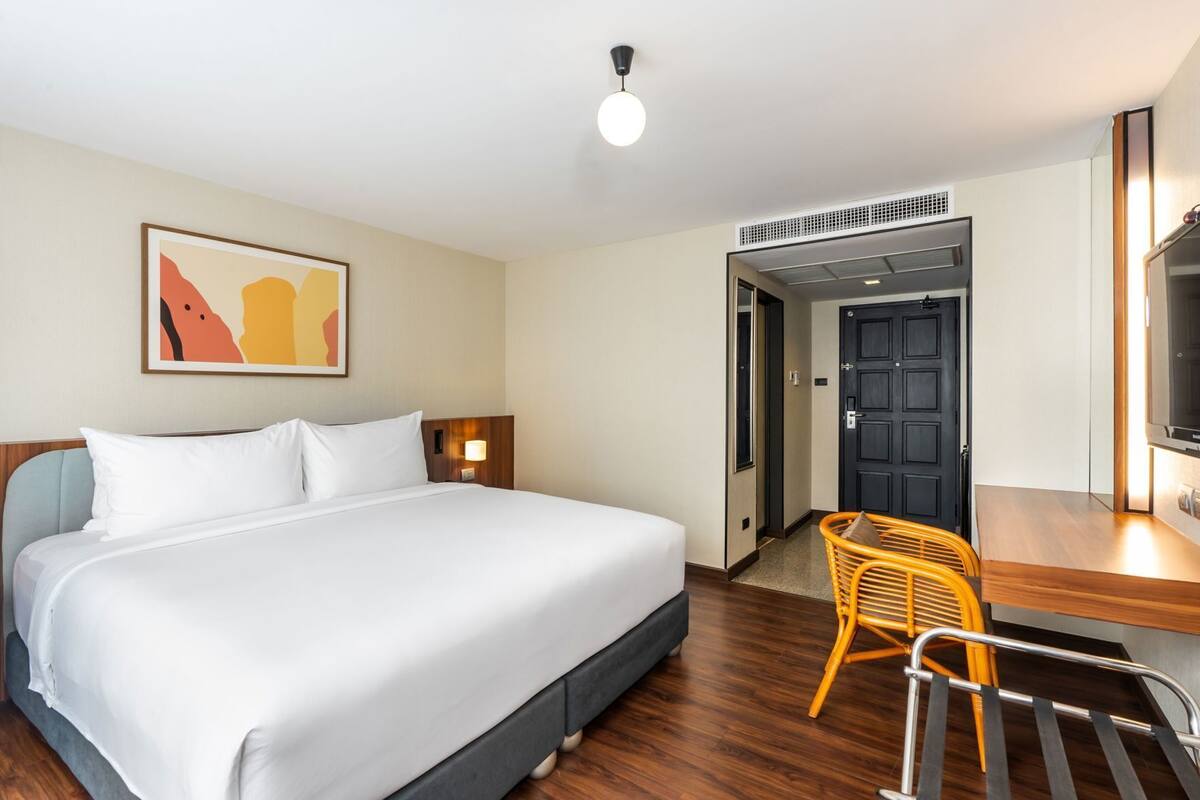 A hotel room with a bed, a desk, a chair, a wall-mounted TV, a door, and a ceiling light.