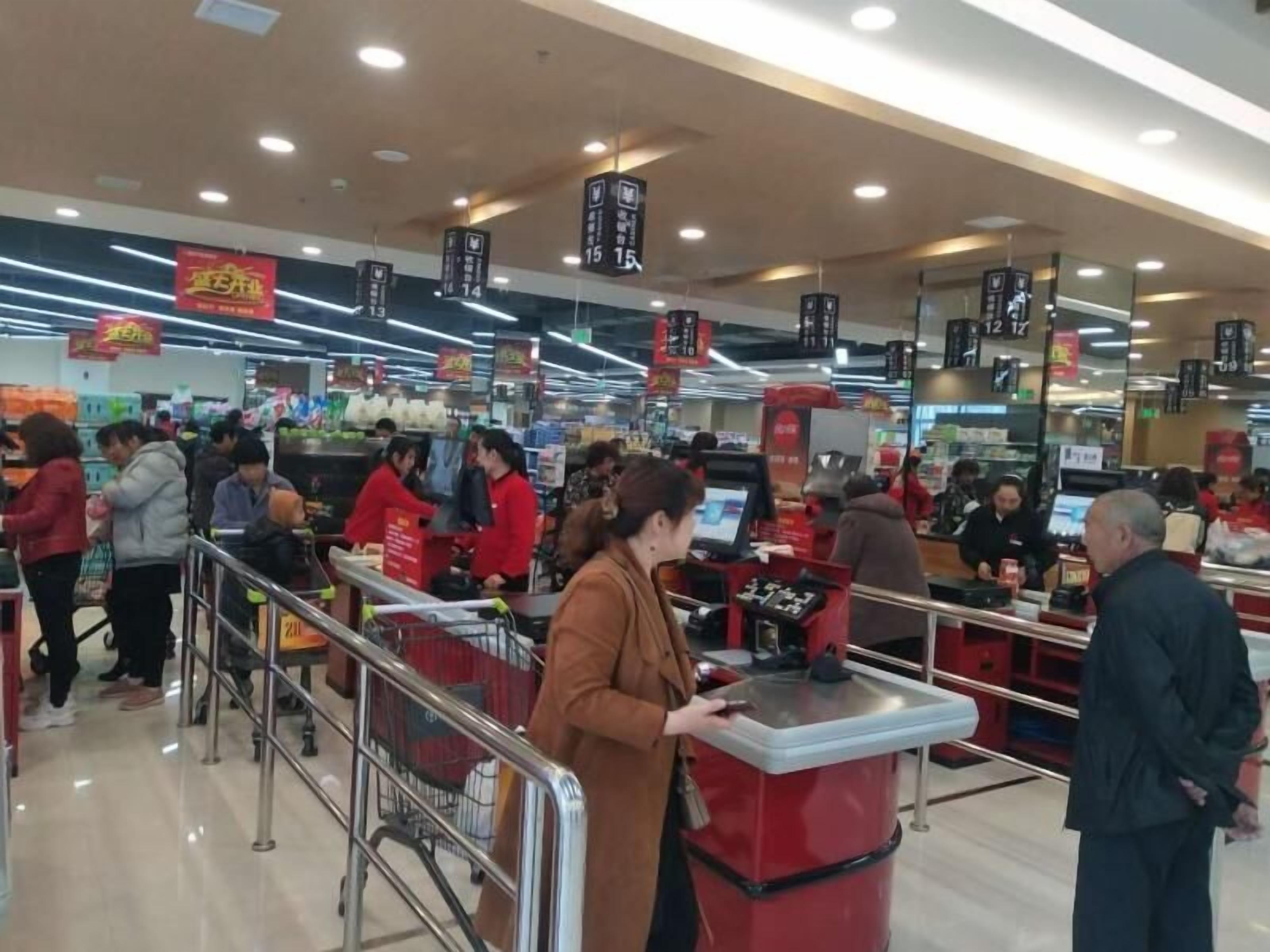 A busy supermarket with shoppers and staff, checkout counters, and promotional signs.