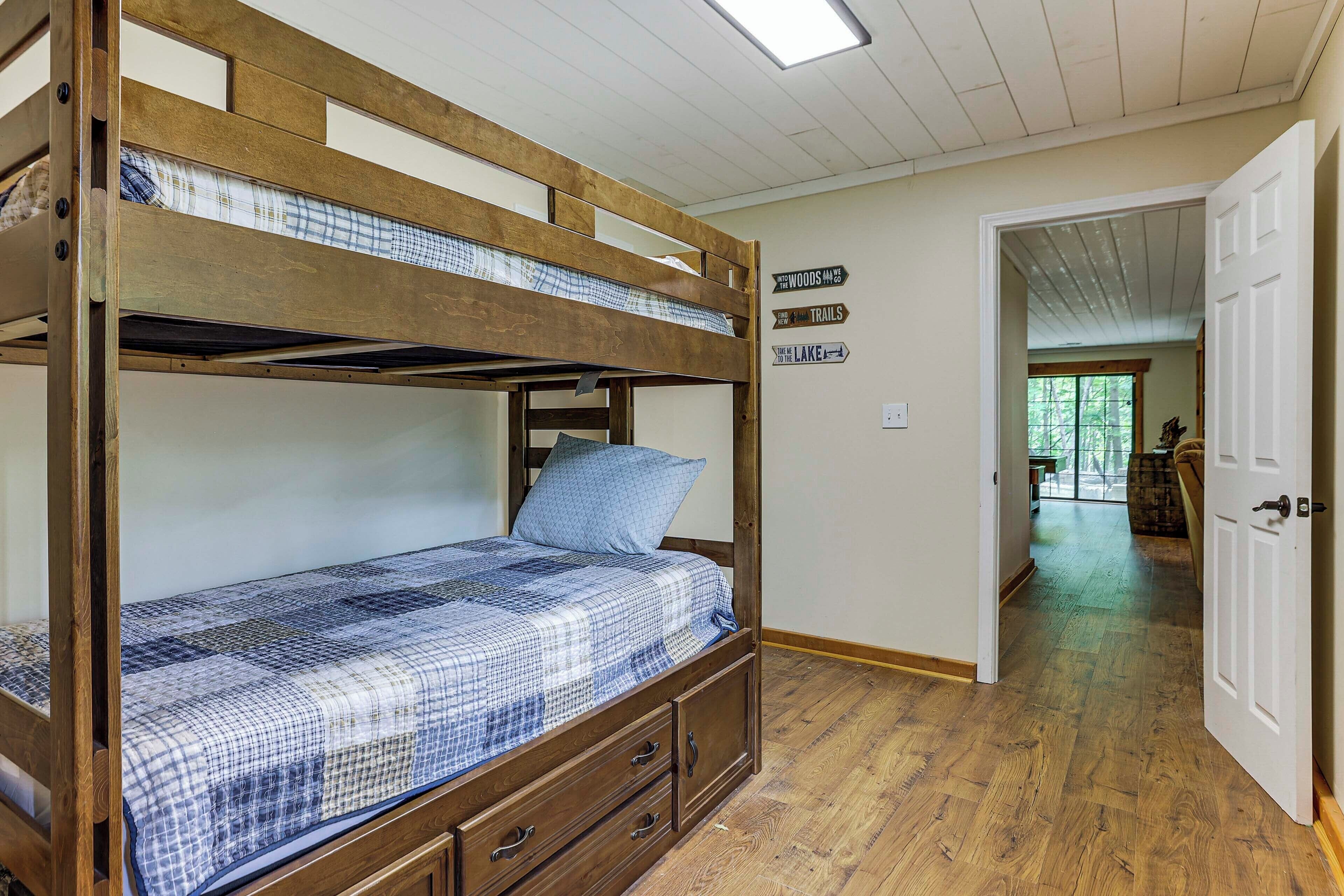 A bunk bed with a checkered bedspread, a pillow, and a small drawer unit. There are signs on the wall indicating directions to 'The Woods' and 'The Lake'.