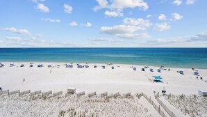 A beach with white sand, clear blue water, and a few people. There are blue umbrellas and beach chairs. - Crystal Shores West 108- The Tide is Calling your Name! Can you Hear it? Book To (Gulf Shores)