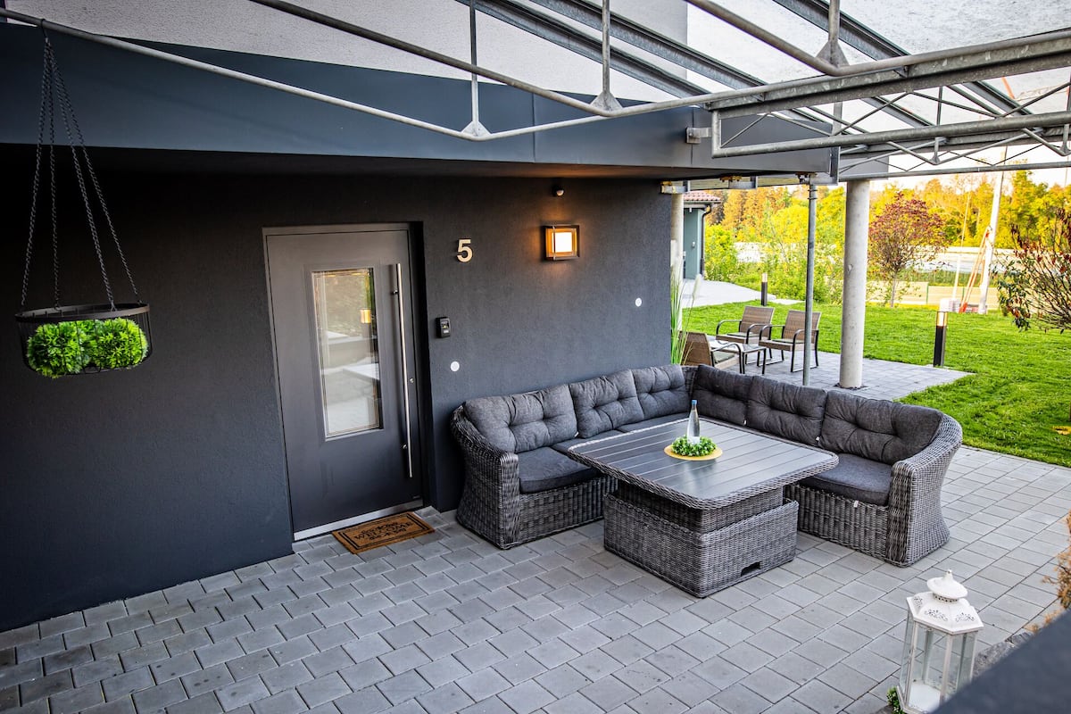 Ein moderner Außenbereich mit einer dunkelgrauen Couch und einem Tisch, einer Tür mit Fensterteil und der Zahl '5' an der Wand.