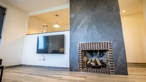 A modern living room with a flat-screen TV mounted on the wall, a fireplace with a brick surround, and a wooden floor. - Familien am Hang (Friedrichshafen)
