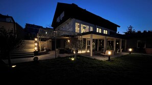 A modern house with a covered patio, outdoor seating, and a well-lit garden area. - Familien am Hang (Friedrichshafen)