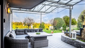 A covered outdoor seating area with a sofa and tables, a pergola structure, and a view of a landscaped garden. - Familien am Hang (Friedrichshafen)