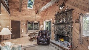 A cozy cabin interior with a stone fireplace, wooden walls, a leather armchair, and a dining area. - Rock'inn Pines by Ruidoso Vacation Rentals (Ruidoso)