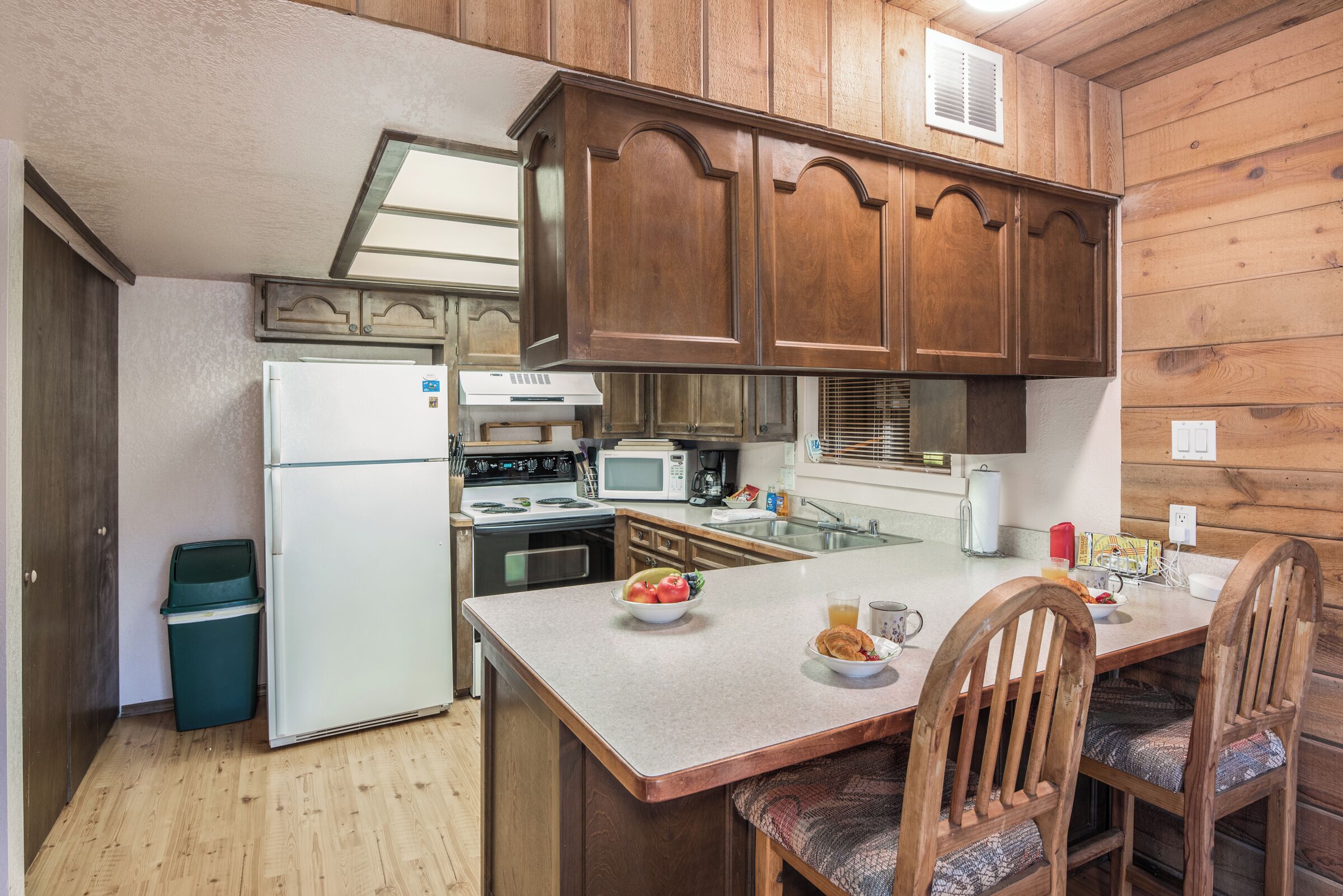 A compact kitchen with wooden cabinets, a refrigerator, microwave, stove, and a dining area with chairs.
