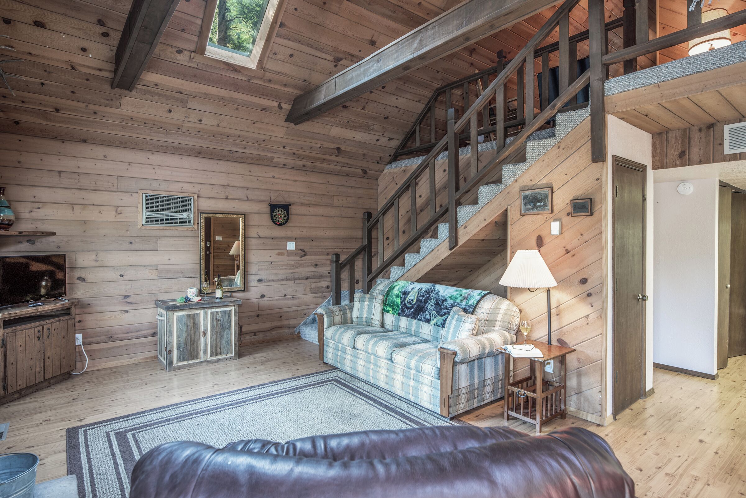 Interior de una cabaña de madera con una escalera, un sofá a cuadros, un reposapiés de cuero y una mesita de noche con lámpara.