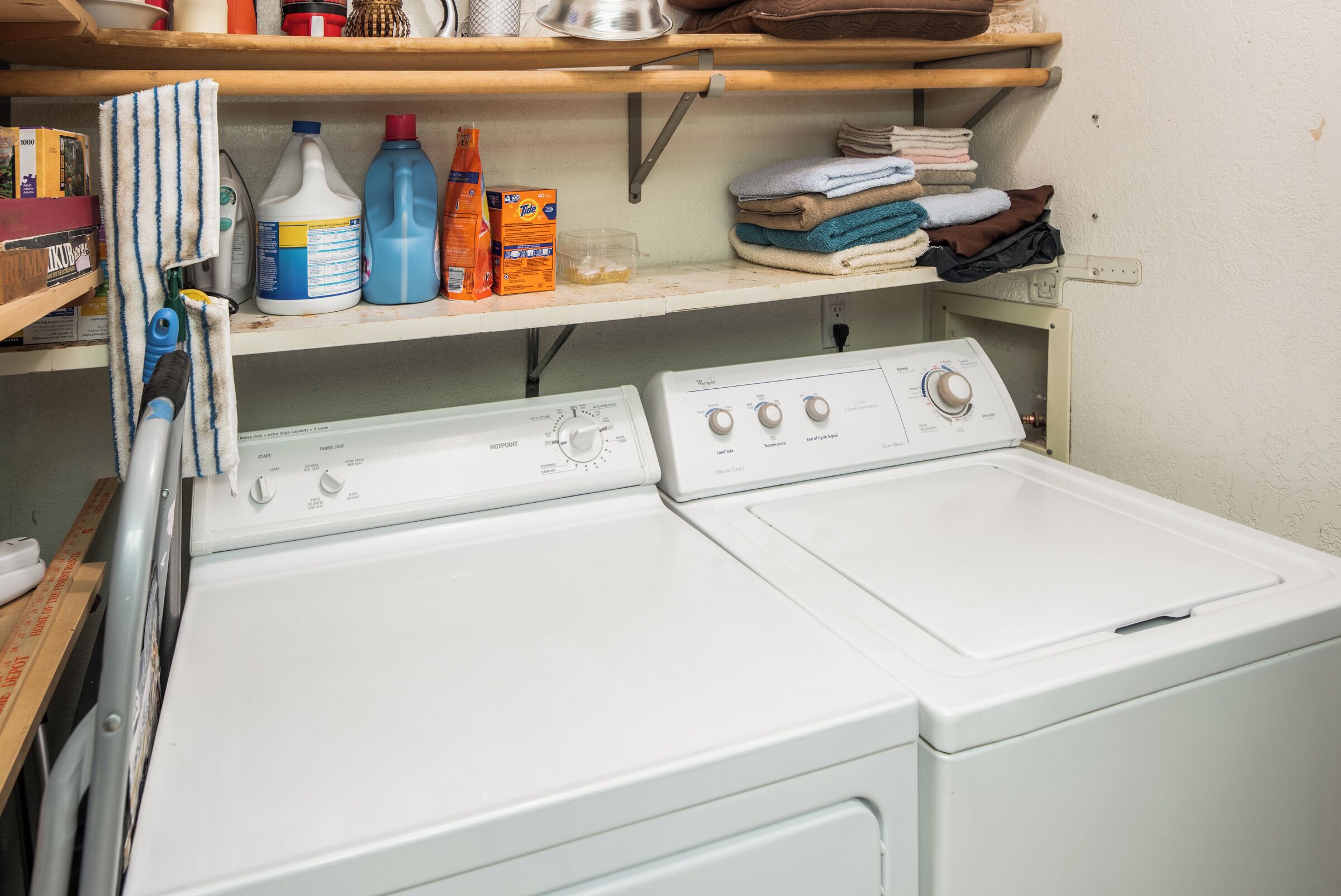 A laundry area with two white front-loading washing machines, shelves with cleaning supplies, and folded towels.