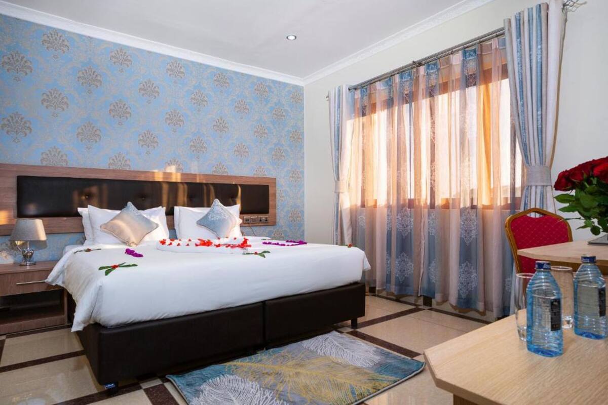A hotel room with a bed, a nightstand, a table with water bottles, and a chair.