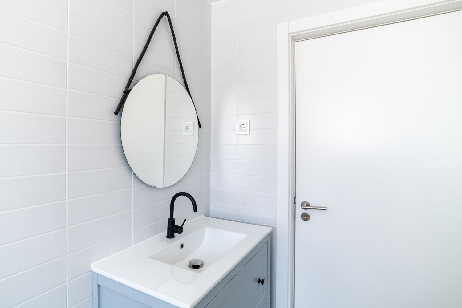 Een badkamer met witte betegelde wanden, een ronde spiegel, een witte wastafel met een zwarte kraan, en een deur met een zilveren handgreep.