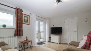 Uma sala de estar com sofá, televisão, uma mesinha com chaleira e uma janela com cortinas.