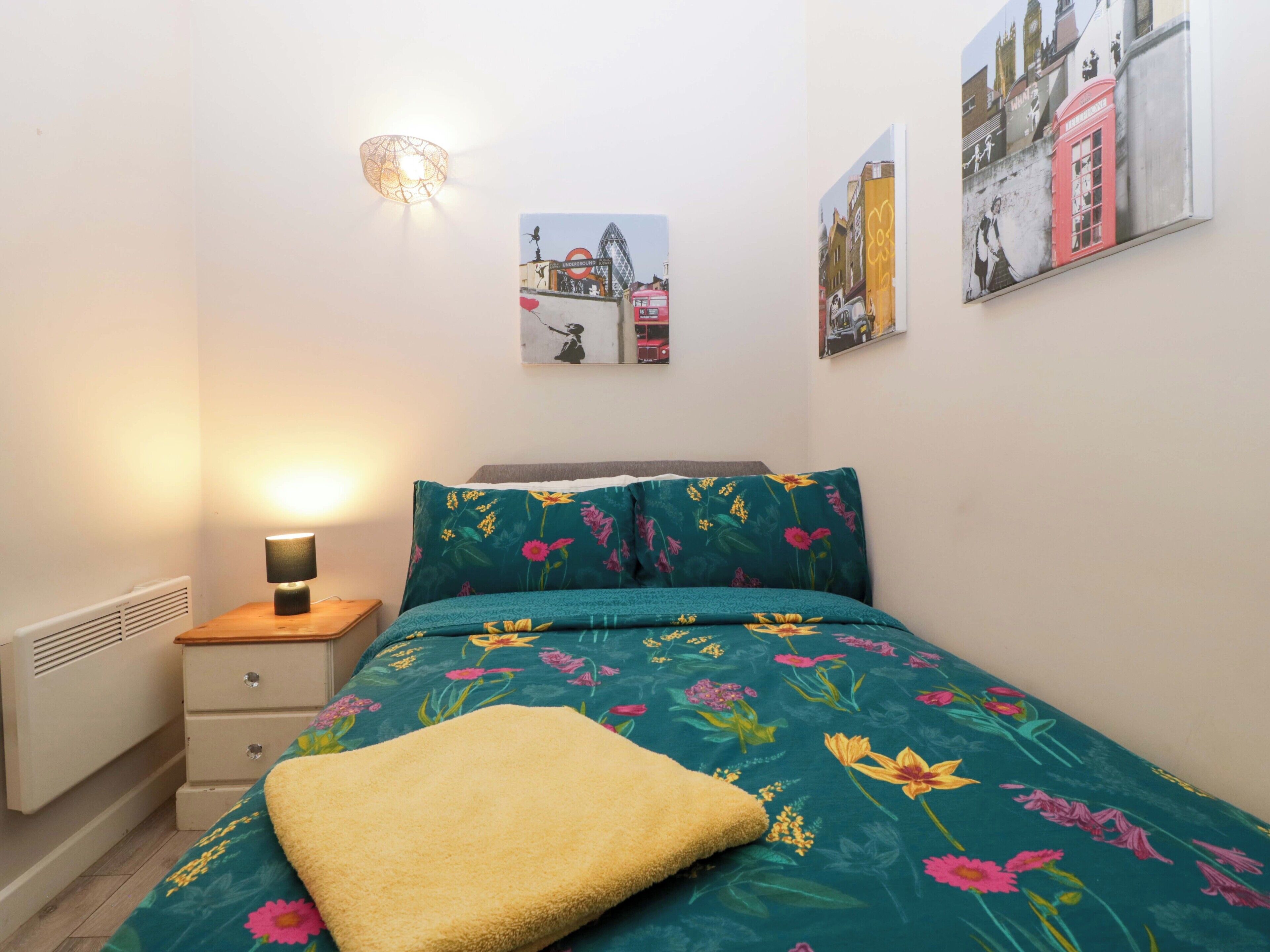A bedroom with a bed, a bedside table, a lamp, and a radiator. There are two wall-mounted pictures depicting city scenes.