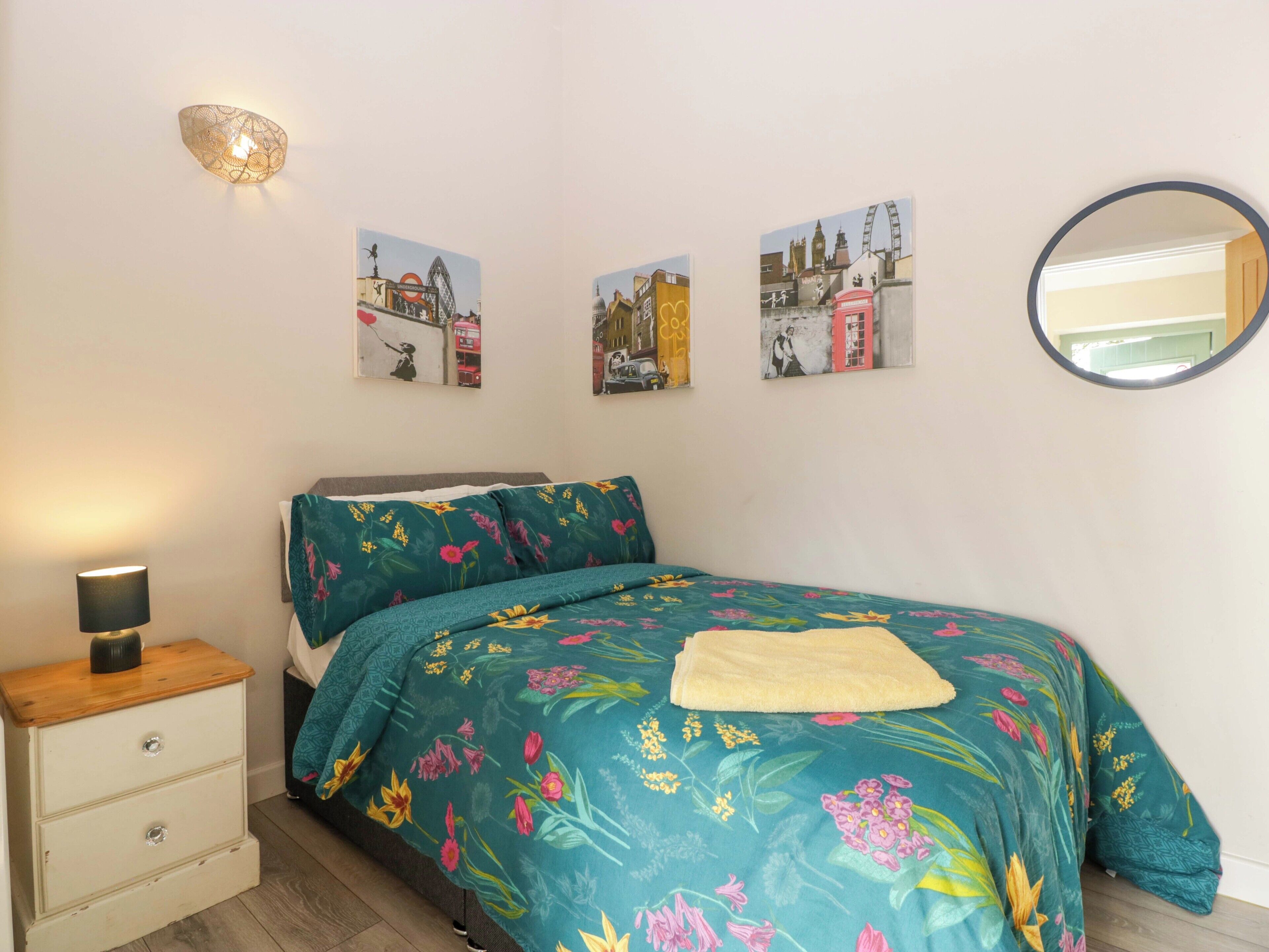 A bedroom with a floral bedspread, a bedside lamp, a dresser, and three wall-mounted pictures depicting cityscapes.