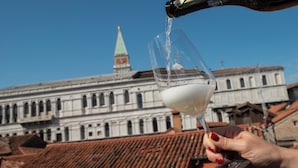 Une main tenant un verre à vin dans lequel on verse du vin à partir d’une bouteille, avec un bâtiment historique en arrière-plan.