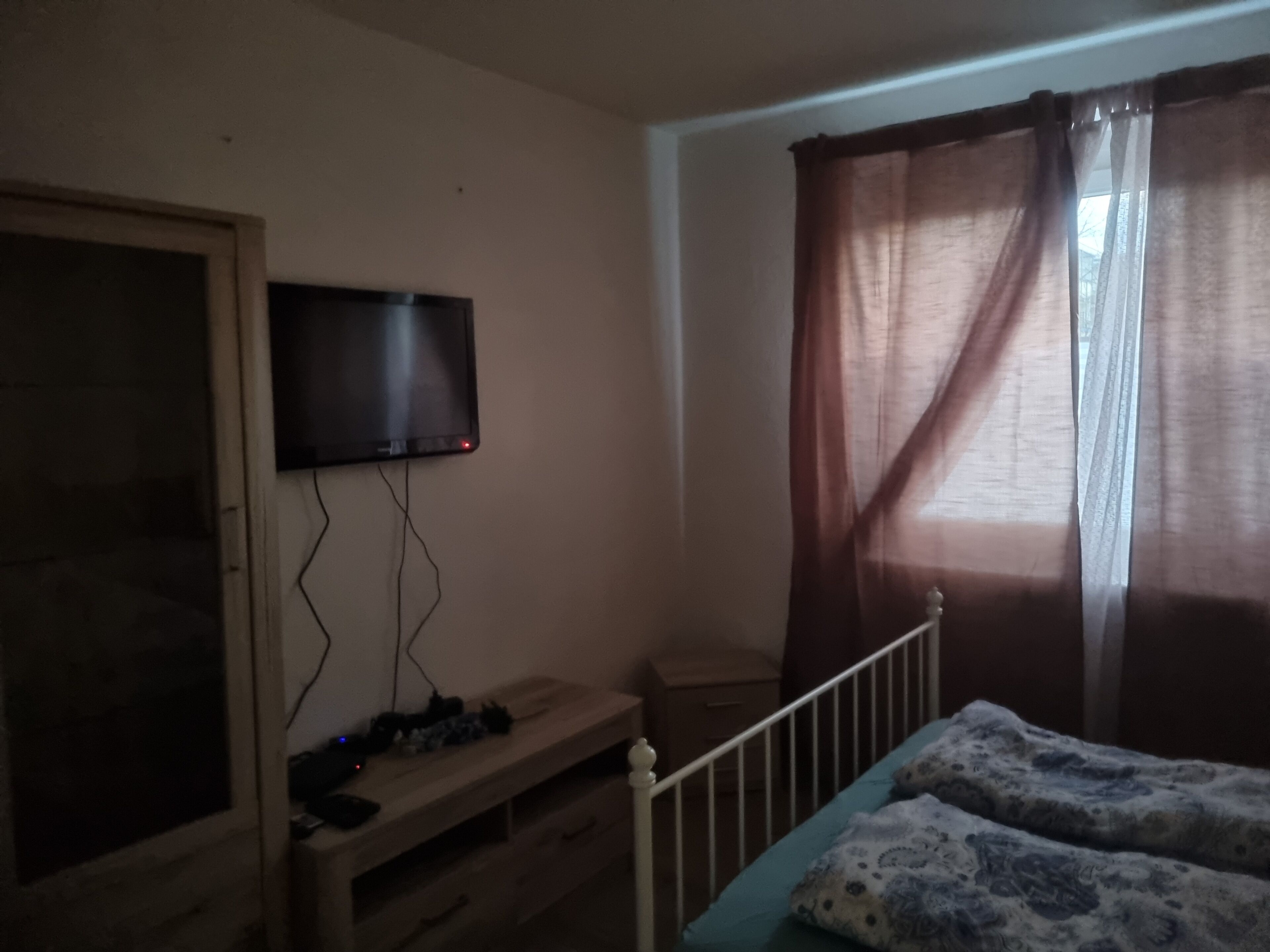 Un dormitorio con una cama, un televisor montado en la pared, un aparador de madera y una ventana con cortinas.