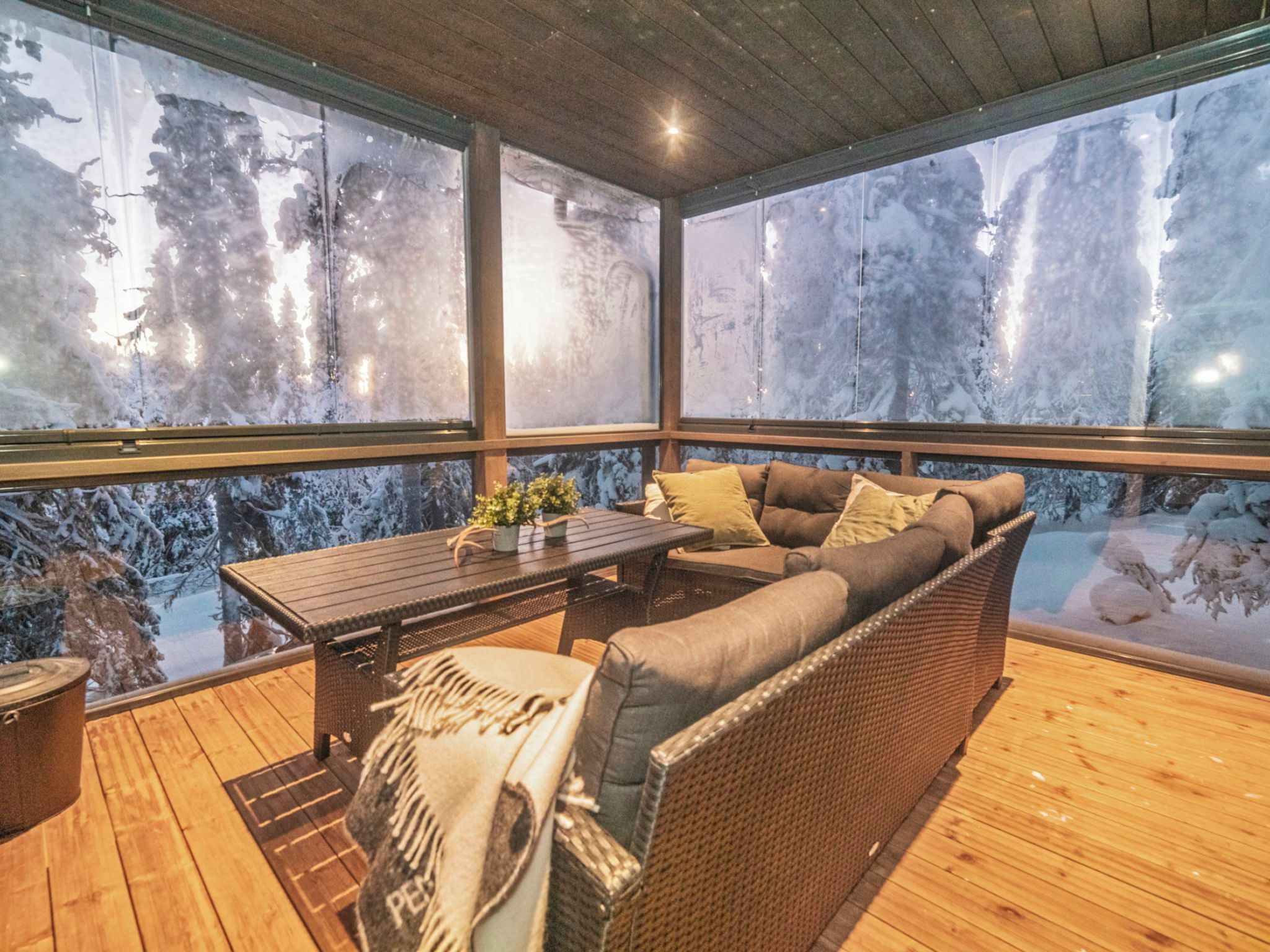 A covered outdoor seating area with a wooden table, cushioned seating, and a view of snow-covered trees.
