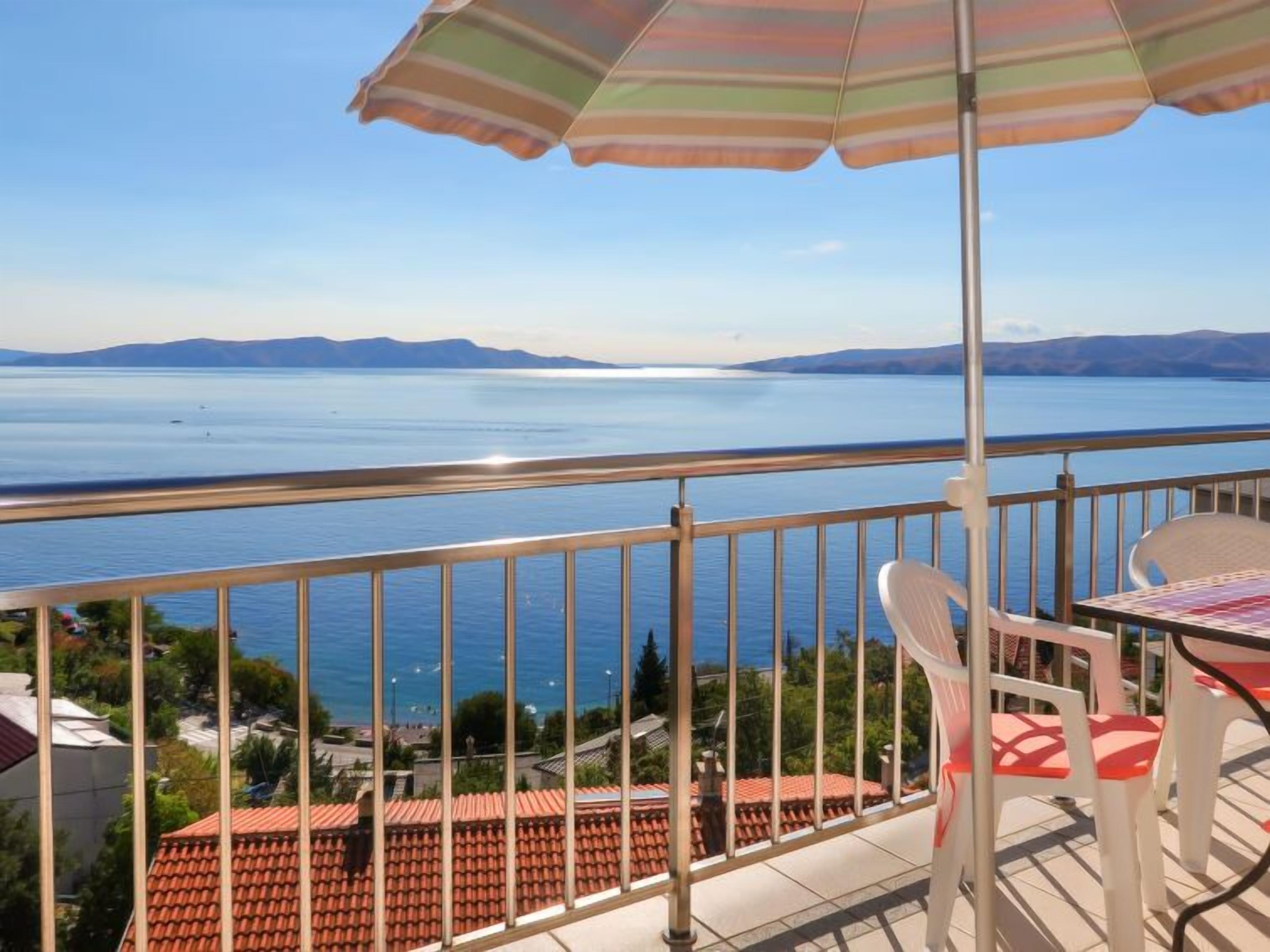 A balcony with a view of a lake, a table with chairs, and a striped umbrella.