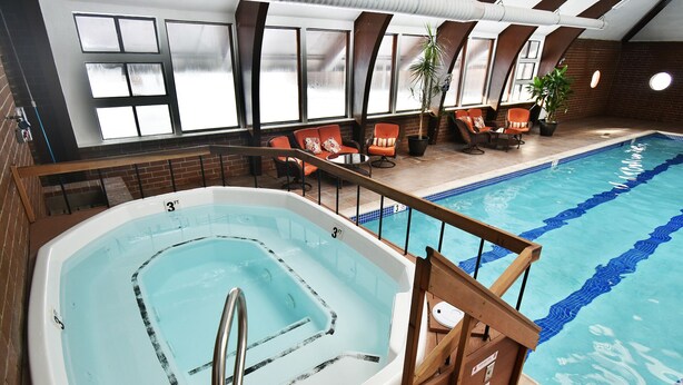 An indoor swimming pool with a hot tub, wooden deck, and seating area with chairs and potted plants.