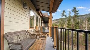 A wooden deck with wicker furniture, a table, and a railing overlooking a forested area.