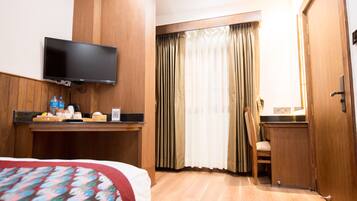 Quarto de hotel com cama, televisão na parede, escrivaninha com cadeira e janela com cortinas.