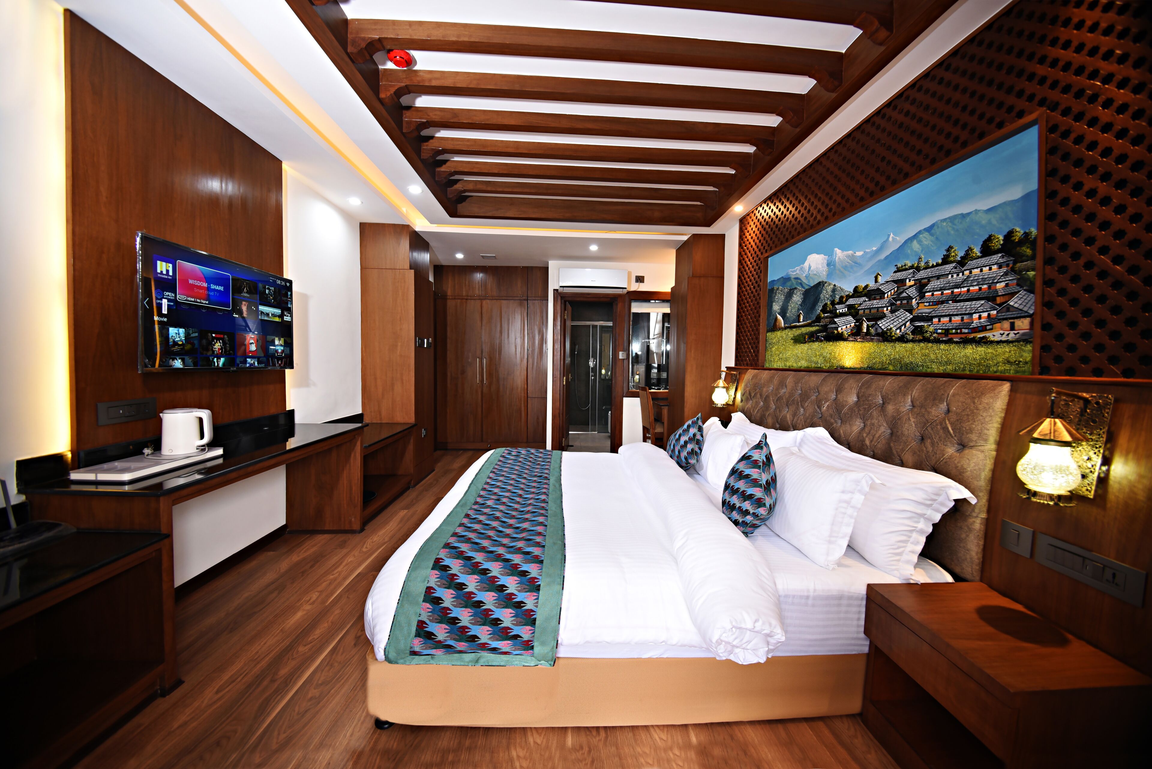 A modern hotel room with a large bed, a flat-screen TV, wooden paneling, and a painting of a mountain landscape.