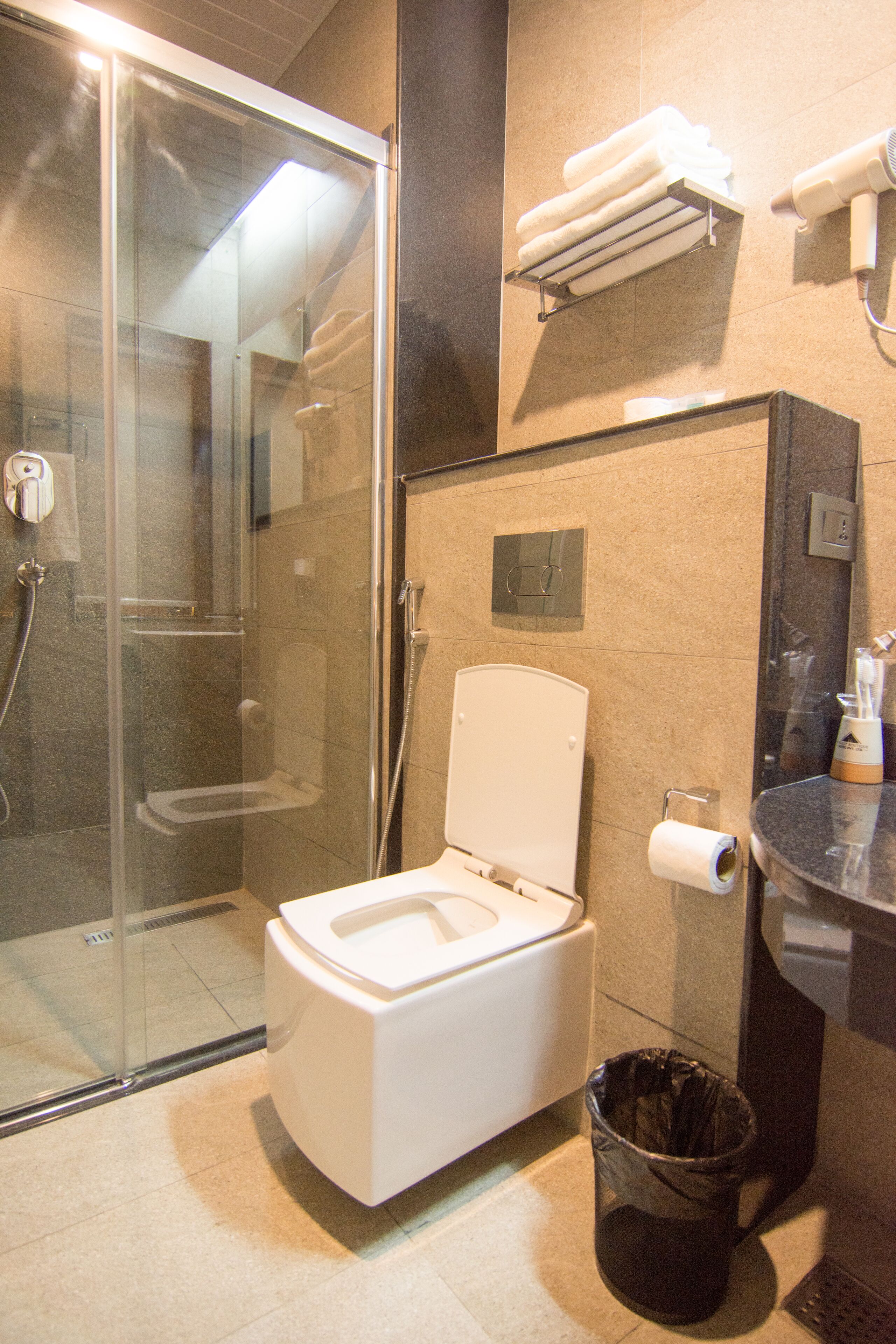 A modern bathroom with a glass-enclosed shower, a toilet, a trash can, and a towel rack with folded towels.