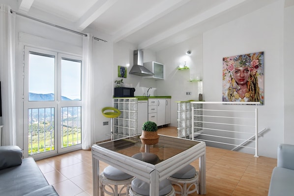 A modern kitchen with a large window, a dining area with a glass table, and a painting on the wall.