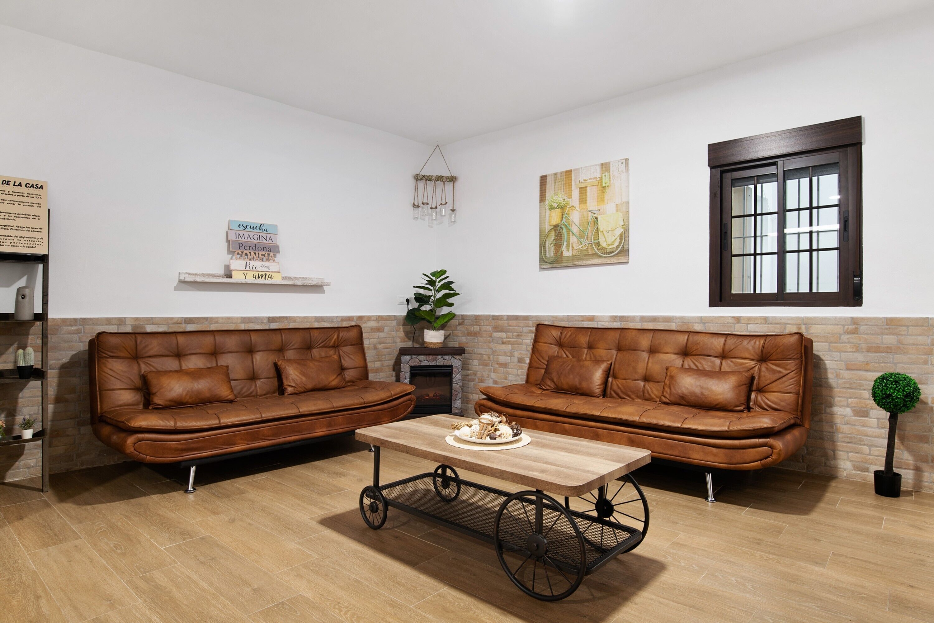 Un salon avec deux canapés en cuir marron, une table basse en bois, une cheminée, une étagère fixée au mur et une fenêtre à cadre sombre.