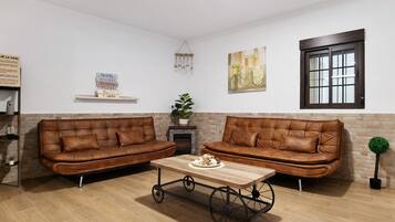 Un salon avec deux canapés en cuir marron, une table basse en bois, une cheminée, une étagère fixée au mur et une fenêtre à cadre sombre.