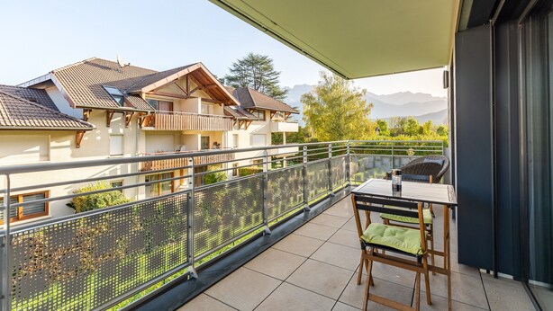 A balcony with a table and chairs, a view of neighboring houses, and a mountain backdrop. - 'Grand Studio by the Lake' with Mountain Views, Terrace and Wi-Fi. (Saint-Jorioz)
