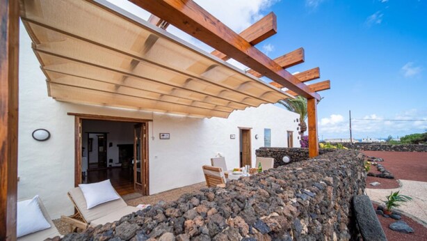 Een gebouw met stenen muren, een houten pergola, een lounge stoel en een tafel met een fles.