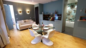 Ein modernes Wohnzimmer mit einem Glastisch, weißen Stühlen, einem beigen Sofa und einer Küchenzeile mit grauen Schränken und einer schwarzen Arbeitsplatte.