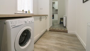 A compact laundry room with a front-loading washing machine, wooden countertop, and a view into the kitchen area. - Rockstar Cottage (Macduff)