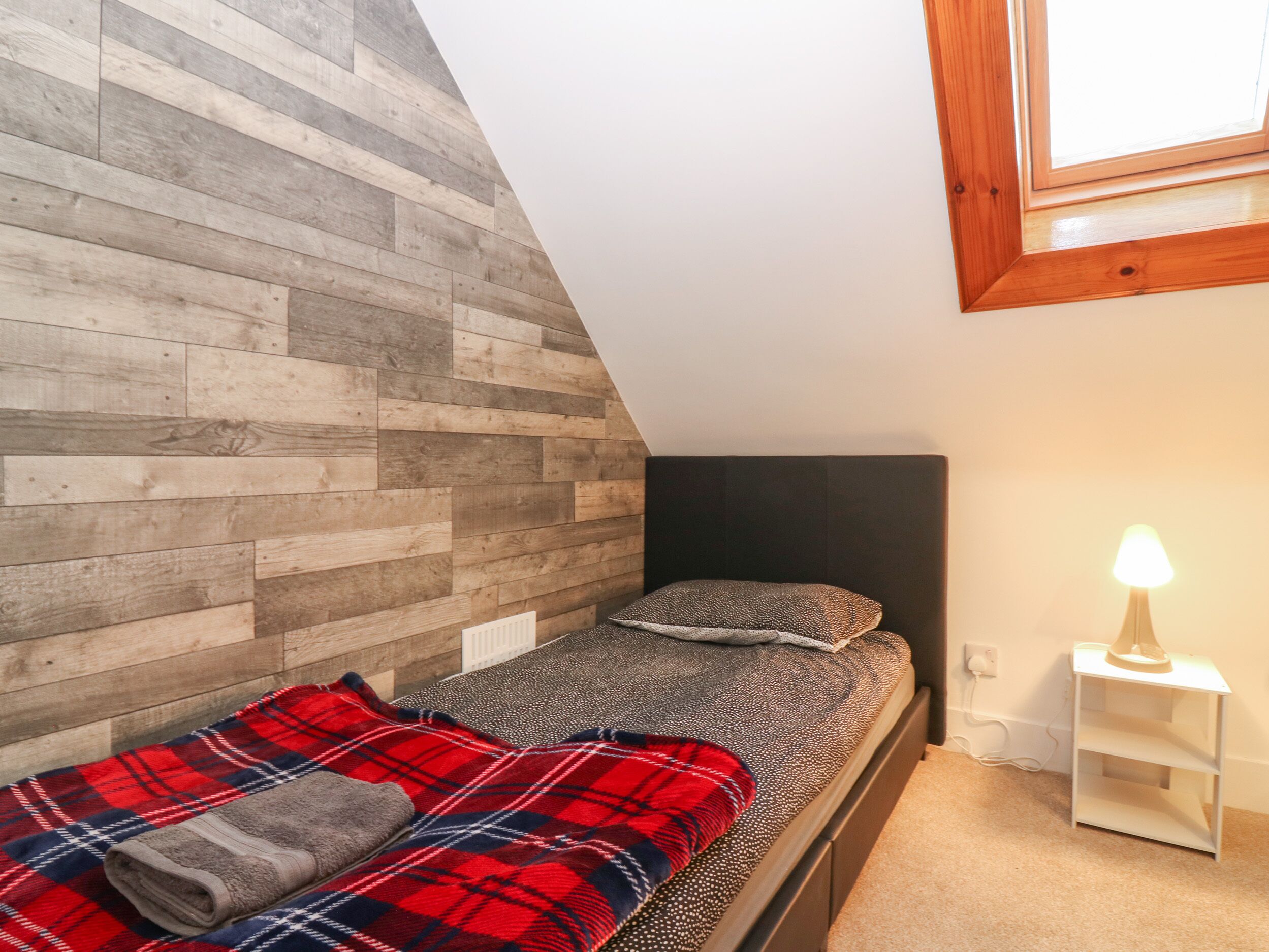 A small bedroom with a sloped ceiling, a wooden wall, a bed with a red and blue plaid blanket, a pillow, and a bedside table with a lamp.