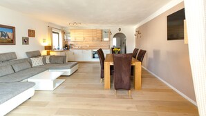 A modern living room with a sectional sofa, a dining table with chairs, and a kitchen area with wooden cabinets and a built-in oven.