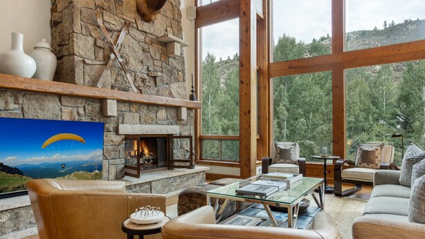 A cozy living room with a stone fireplace, leather sofas, and a large window offering a forest view.