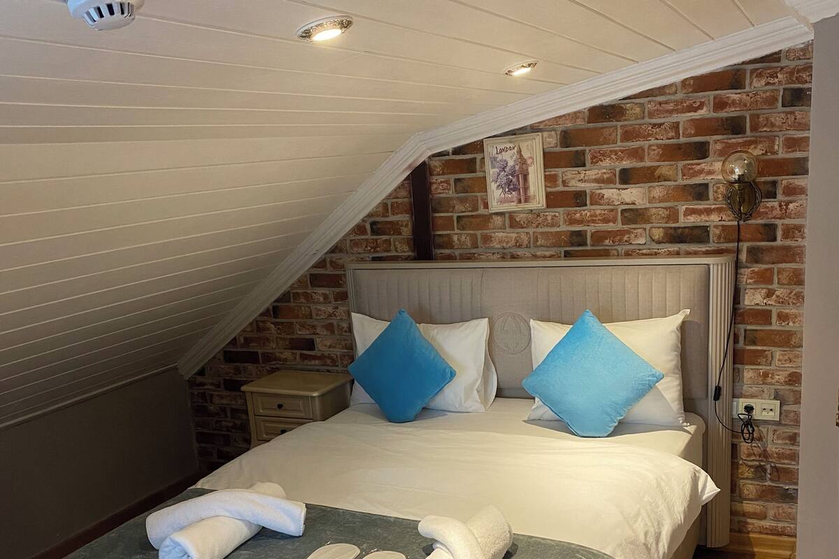 A neatly made bed with white linens and blue pillows, a small bedside table, and a brick wall with a framed picture.