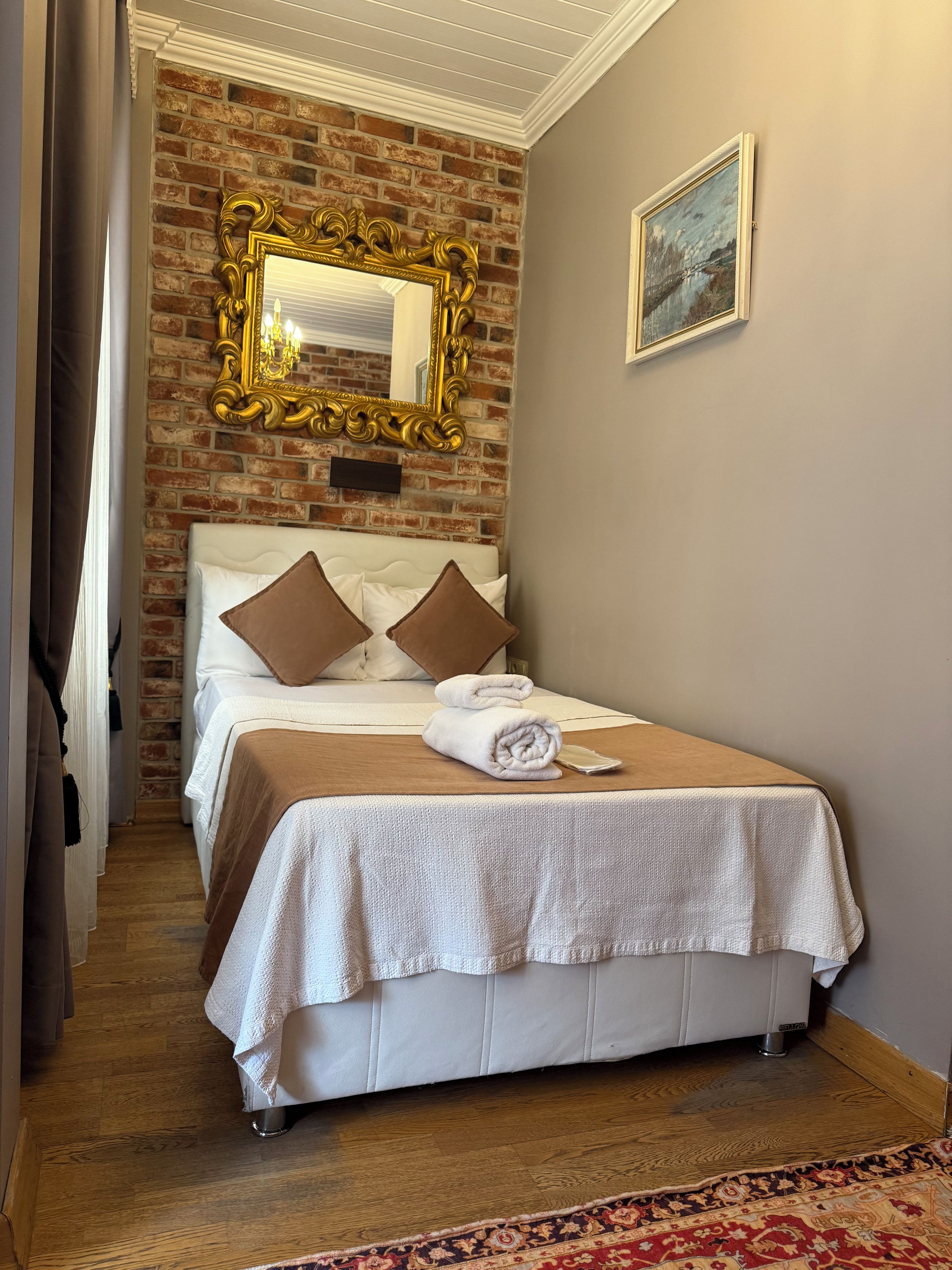 Una habitación de hotel con una pared de ladrillo, un espejo grande y ornamentado, una cama con ropa blanca y almohadas marrones, y un cuadro en la pared.