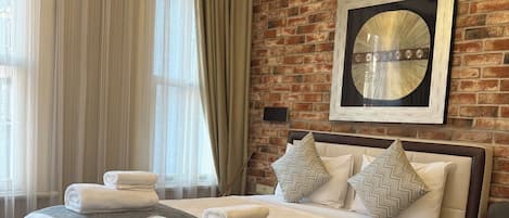 A hotel room with a large bed, a chandelier, a brick wall, and a large window with curtains.