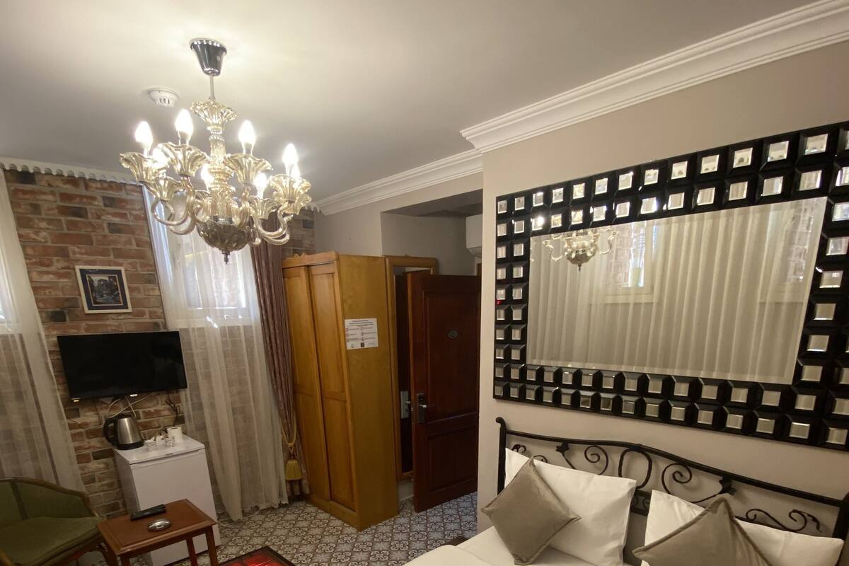 A hotel room with a bed, a TV, a wooden wardrobe, a desk with a chair, and a chandelier.