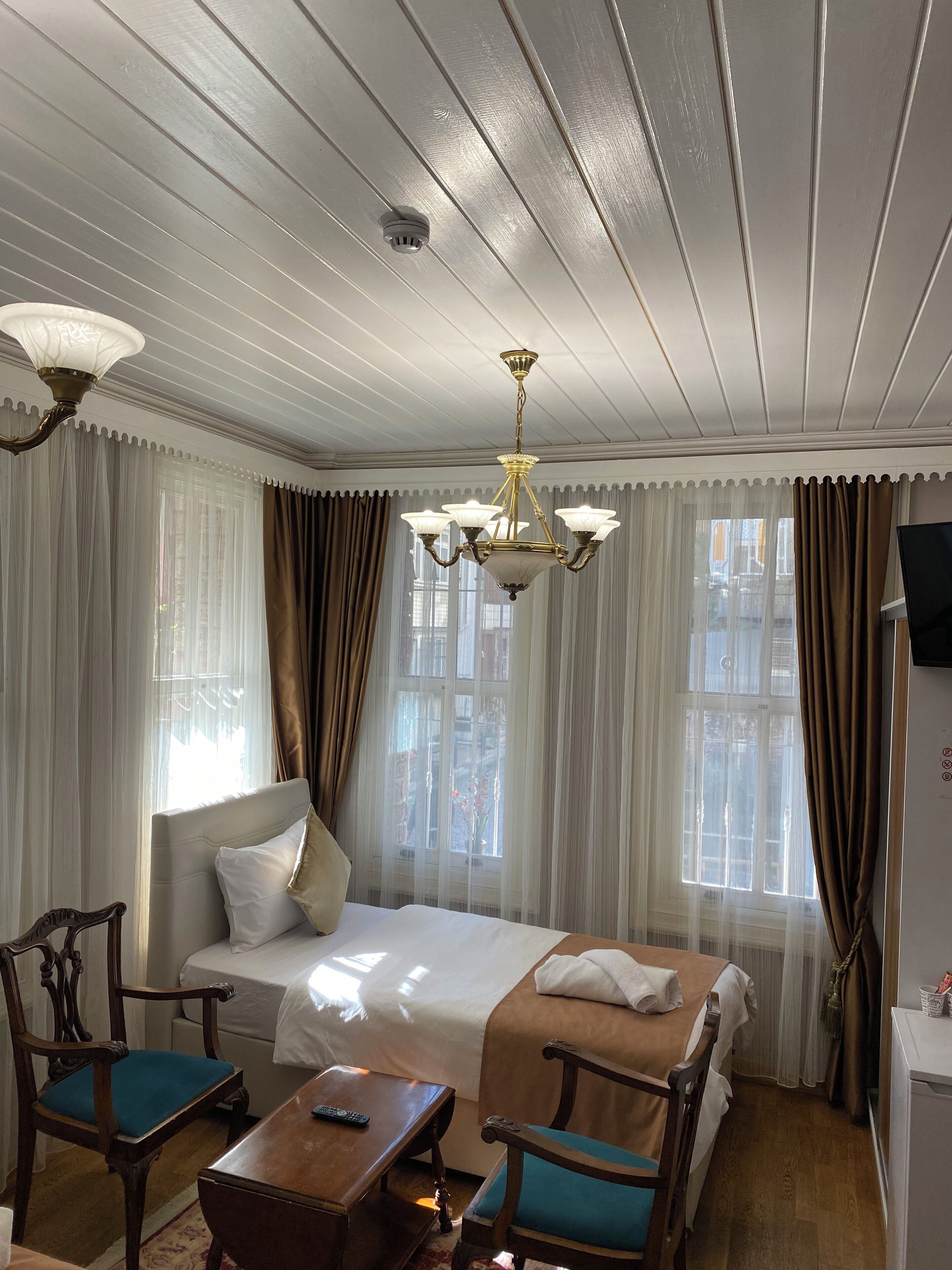 A hotel room with a bed, two chairs, a small table, a chandelier, and a television on the wall.