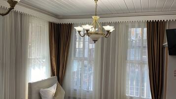 A hotel room with a bed, two chairs, a small table, a chandelier, and a television on the wall.