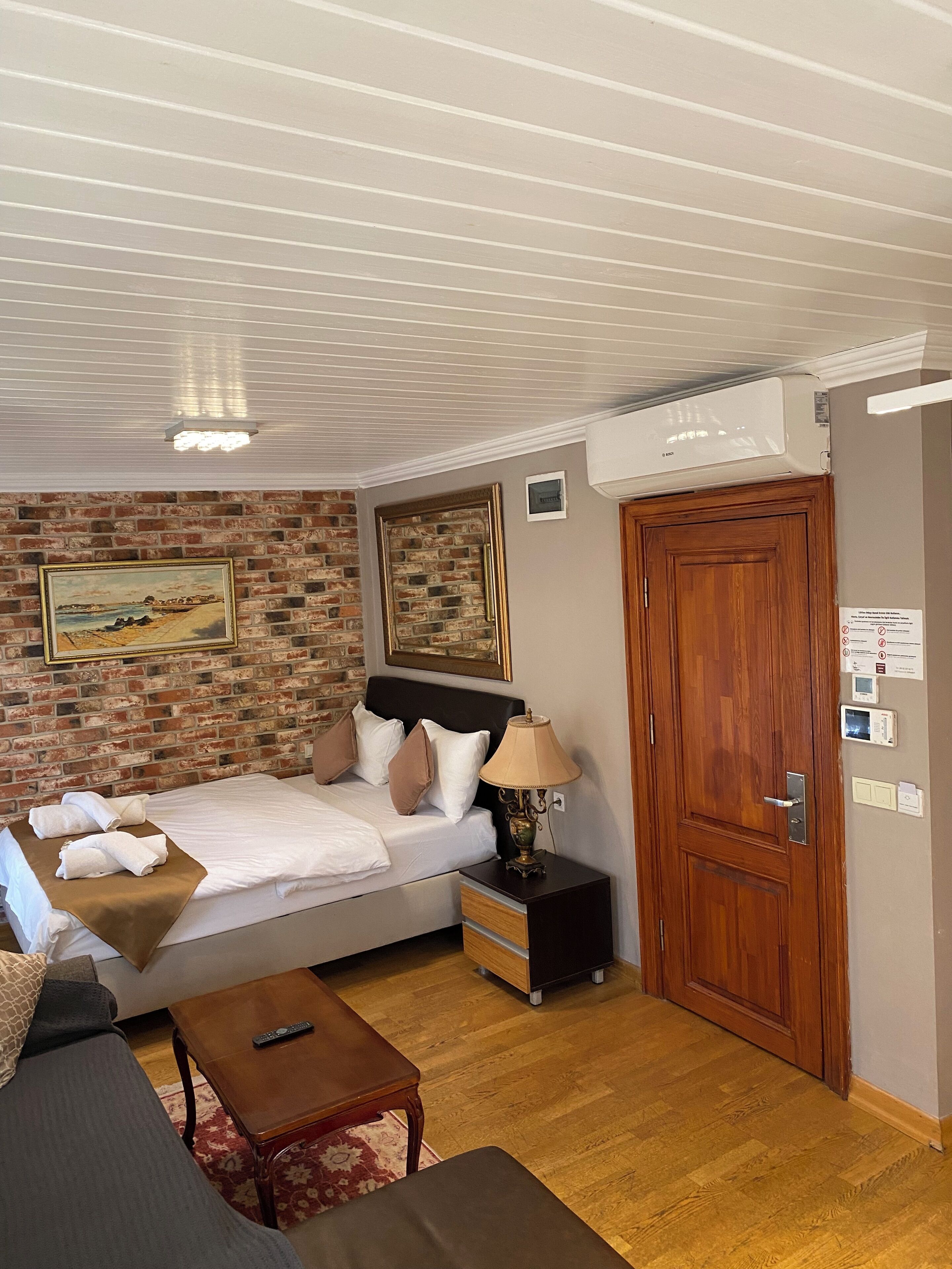 A bedroom with a brick wall, a bed with pillows, a bedside table with a lamp, a wooden door, and an air conditioning unit.